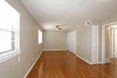 This single-floor space is characterized by warm, polished hardwood flooring that runs throughout the main living and bedroom areas, lending a cohesive and classic look to the interior. The neutral greige wall tones complement the natural wood tones, creating a clean and versatile aesthetic.

The spacious main living area benefits from multiple windows fitted with white blinds, allowing ample natural light to fill the room. A ceiling fan is installed overhead, adding both functionality and airflow. Built-in shelving is visible along one wall, offering practical storage options.

The bedroom features its own set of windows for natural light, hardwood flooring consistent with the rest of the unit, and a double-door closet providing generous storage. The layout connects efficiently to the rest of the space through a central hallway.

The kitchen is equipped with white cabinetry, laminate countertops, a freestanding electric range, and tile flooring with a wood-look pattern. A window above the counter area adds natural light, and an exterior door provides direct outside access.

The bathroom is neatly appointed with large-format tile flooring in a wood-tone finish, a white vanity with mirror, a tub-shower combination, and a window for ventilation and natural light.