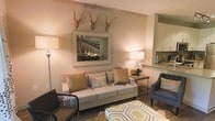 This single-floor space showcases a thoughtfully designed living area that blends rustic and contemporary elements. The living room serves as the focal point, centered around a plush, neutral-toned sofa adorned with textured throw pillows in warm earth tones. A large framed artwork depicting an urban bridge scene anchors the main wall, while a pair of decorative antler mounts above adds a distinctive rustic touch.

The space is warmed by soft ambient lighting provided by floor and table lamps, creating an inviting atmosphere. Dark hardwood-style flooring grounds the room, complemented by a geometric-patterned area rug in muted yellow and white tones. Seating is varied and functional, with additional chairs—including a woven accent chair and an upholstered armchair—offering flexible arrangements.

A side table with a wood-and-metal base provides practical surface space beside the sofa. The living area flows naturally into a kitchen, visible in the background, which features light cabinetry, a tile backsplash, and stone countertops, suggesting an open-concept layout that enhances the overall sense of connectivity and space.