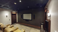 This dedicated home theater is thoughtfully designed to deliver an immersive cinematic experience. The room is anchored by a large wall-mounted flat-panel screen flanked by tall, slim surround sound speakers and a soundbar, ensuring comprehensive audio coverage throughout the space. A ceiling-mounted projector provides an additional viewing option, offering flexibility for different screening preferences.

Seating consists of multiple rows of tan leather recliner chairs, each equipped with integrated cup holders and padded armrests, providing both comfort and practicality during extended viewing sessions. The rows are arranged to offer clear sightlines to the screen from every seat.

The dark drop ceiling with recessed lighting creates a controlled lighting environment ideal for screening, while wall-mounted sconces on the lighter rear and side walls provide soft ambient illumination. Dark, textured wall treatments on the screen wall enhance the theater aesthetic and aid in sound absorption.

Rich wood trim, including a decorative column and door surround, adds warmth and architectural interest to the room. A beverage refrigerator near the entrance adds convenience. A patterned area rug grounds the seating area, complementing the carpeted flooring. The overall ambiance is that of a well-appointed, purpose-built private screening room.