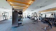 This generously sized fitness center is a standout exercise facility designed to support a full range of workout routines. The space is immediately distinguished by its eye-catching architectural ceiling feature — a sculptural, wave-like wooden lattice installation that runs across much of the ceiling, adding warmth and visual interest to the otherwise clean, modern interior. Multiple large ceiling fans and recessed lighting keep the space bright and comfortable throughout.

The strength training zone is comprehensively outfitted with a Smith machine, a full rack of dumbbells, weight benches, barbells, and a multi-station functional training rig complete with kettlebells, medicine balls, battle ropes, and plyo boxes. A large wall mirror amplifies the sense of space and supports proper form. Wall-mounted TVs provide entertainment throughout the facility.

The cardio section lines the perimeter and includes treadmills, stair climbers, an elliptical, and a Peloton cycle. A heavy punching bag adds a boxing element to the functional fitness offerings. Patterned, low-pile carpet with teal accents covers the floor throughout, giving the space a polished yet athletic feel. Large black-framed windows along one side flood the room with natural light and offer views of an outdoor green space, creating an open, inviting atmosphere conducive to focused, effective training.