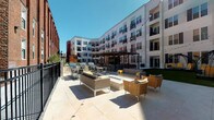This outdoor courtyard and pool area at 2403 7th Boulevard in St. Louis, Missouri, is a thoughtfully designed amenity space nestled within a contemporary multi-story residential building. The courtyard is framed on multiple sides by the building's modern white facade, accented with dark shutters and private balconies, creating an intimate yet open-air atmosphere bathed in natural sunlight.

The hardscaped patio features large-format light stone tile flooring that extends generously across the central gathering area. Comfortable lounge seating, including woven accent chairs and neutral-toned sofas with bold yellow accent pillows, is arranged around a central fire table, creating an inviting social hub. String lights drape overhead, adding ambiance during evening hours.

A pergola-covered zone provides shaded seating and houses multiple grilling stations alongside high-top dining tables, ideal for outdoor cooking and casual entertaining. A section of manicured artificial turf adds a green contrast, and distinctive dome-shaped pod swings offer unique relaxation spots. Secured black iron fencing and gated access points define the perimeter, ensuring privacy. The entry passage features clean architectural detailing with dark-framed glass doors and decorative planters.