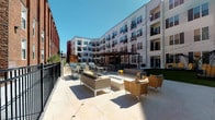 This outdoor courtyard and pool area at 2403 7th Boulevard in St. Louis, Missouri, is a thoughtfully designed amenity space nestled within a contemporary multi-story residential building. The courtyard is framed on multiple sides by the building's modern white facade, accented with dark shutters and private balconies, creating an intimate yet open-air atmosphere bathed in natural sunlight.

The hardscaped patio features large-format light stone tile flooring that extends generously across the central gathering area. Comfortable lounge seating, including woven accent chairs and neutral-toned sofas with bold yellow accent pillows, is arranged around a central fire table, creating an inviting social hub. String lights drape overhead, adding ambiance during evening hours.

A pergola-covered zone provides shaded seating and houses multiple grilling stations alongside high-top dining tables, ideal for outdoor cooking and casual entertaining. A section of manicured artificial turf adds a green contrast, and distinctive dome-shaped pod swings offer unique relaxation spots. Secured black iron fencing and gated access points define the perimeter, ensuring privacy. The entry passage features clean architectural detailing with dark-framed glass doors and decorative planters.