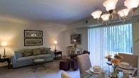 This single-floor residence presents an open-concept layout that seamlessly connects the living and dining areas into one cohesive, functional space. The living area is finished with plush wall-to-wall carpeting and features a comfortable sofa with decorative accent pillows, an armchair, and an upholstered ottoman, all arranged around a glass-top coffee table with ornate metal detailing. Framed artwork adorns the walls, and a classic table lamp adds a warm ambient glow that complements the room's traditional aesthetic. A white storage cabinet serves as a decorative accent piece, adding both style and practicality. A large sliding glass door or window fitted with vertical blinds allows generous natural light to fill the space, offering a view of greenery outside. The dining area is anchored by a glass-top table paired with upholstered chairs and illuminated by an elegant chandelier with multiple globe lights. The overall ambiance is warm and inviting, with traditional furnishings and neutral tones creating a comfortable, well-appointed living environment throughout.