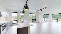 This contemporary single-floor space offers a thoughtfully designed open-concept layout that seamlessly connects the kitchen, dining, and living areas. The kitchen is a standout feature, showcasing two-tone cabinetry with gray lower cabinets and white upper cabinets, a white subway tile backsplash, stainless steel appliances including a refrigerator, range, and microwave, and a generously sized island with a white quartz countertop and an integrated double sink — all illuminated by striking black pendant lights with gold interiors. The open living area extends from the kitchen and is bathed in natural light through an array of large black-framed windows and a sliding glass door that leads to an outdoor balcony, offering views of lush greenery. Light wood-look flooring runs throughout the main living spaces, adding warmth to the modern aesthetic. The bedroom features plush carpeting and corner windows that frame peaceful outdoor views. The bathrooms are clean and functional, each appointed with gray floating vanities, white countertops, and a frameless walk-in shower. Track lighting throughout the unit provides flexible, modern illumination, completing the sleek and cohesive design of this well-appointed space.
