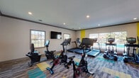 Located at 11601 Nansemond Dr in Louisville, KY, this fitness center is a thoughtfully designed, single-floor facility offering a comprehensive workout experience. The space is organized into distinct exercise zones, each catering to different training needs.

The cardio area is generously outfitted with multiple TRUE-brand treadmills and elliptical machines positioned along large windows that invite abundant natural light and offer views of the surrounding outdoor area, including a neighboring pool. A wall-mounted flat-screen TV enhances the cardio experience.

The cycling zone features several stationary bikes with integrated digital displays, set atop a vibrant patchwork carpet in shades of gray, teal, and yellow — a design element that adds energy and visual interest throughout the space.

A dedicated strength training room houses a multi-station cable machine, an adjustable weight bench, and a dumbbell rack, complemented by a large wall mirror and resistance band station mounted on a bold yellow accent wall. An additional open area provides space for functional fitness with stability balls and wall-mounted equipment for stretching and bodyweight exercises.

Recessed lighting throughout ensures consistent illumination, while crown molding and clean, neutral-toned walls contribute a polished, well-maintained aesthetic. The entrance features hard-surface flooring that transitions smoothly into the carpeted workout zones, creating a welcoming and practical flow through the facility.