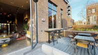 This single-floor space is purpose-built as a professional pet grooming facility, combining functional interior workspaces with an inviting outdoor area. The exterior features classic exposed brick construction with large floor-to-ceiling windows, and a wood-decked terrace furnished with bar-height seating, offering a pleasant outdoor environment adjacent to the main interior.

Inside, the grooming area is defined by white subway-tiled walls and slate-gray floor tiles that establish a clean, clinical aesthetic throughout. A dedicated wash station room houses a row of three stainless steel elevated bathing tubs, each equipped with hose attachments and individual overhead industrial-style pendant lighting mounted against the dark ceiling. Step platforms provide easy pet access to each tub, and a floor drain ensures efficient water management.

A separate grooming room is equipped with a hydraulic lift grooming table, stainless steel utility tables on casters, a wall-mounted professional dryer with a flexible hose, and dedicated HVAC equipment to maintain proper ventilation. Branded wall graphics, including a playful logo and the phrase "Live Like Someone Left the Gate Open," add personality to the space. A glass door connects directly to an adjacent area, enhancing workflow and accessibility throughout the facility.