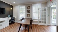 Located at 325 Marymeade Dr in Summerville, SC, this single-floor space combines functional design with refined aesthetics. Warm hardwood flooring runs throughout, tying each area together cohesively. Crown molding adds a classic architectural touch to the ceilings, enhancing the overall polish of the interior.

The co-working area serves as a central hub, featuring a built-in white desk station accommodating multiple workspaces, complete with cabinetry and a wall-mounted flat-screen display set against a bold dark accent wall. Pendant lighting casts a warm glow over the workstations, while a large window invites ample natural light. A rustic wood conference table with studded leather chairs occupies a dedicated meeting zone, accompanied by French glass doors that lend an open, airy quality to the space. Decorative wall art in natural, organic textures adds visual interest.

A welcoming entry area features curated décor, including a round mirror, accent lamp, and abstract wall art, creating a refined first impression. Adjacent to this is a dedicated package room housing smart parcel lockers, offering a practical and secure solution for deliveries. Together, these spaces reflect a well-appointed, community-oriented environment designed for both productivity and convenience.