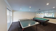 This single-floor game room is designed as a dedicated recreational space, offering a well-appointed layout for leisure and entertainment. The room is anchored by two central game tables — a full-size pool table with a green felt surface, illuminated by an elegant wrought-iron pendant light fixture, and a regulation-size ping-pong table positioned to allow ample play space on all sides. A large wall mirror adjacent to the pool table visually expands the room and reflects natural light throughout the space.

Dark, textured wall-to-wall carpeting covers the floor, providing a clean contrast to the warm beige walls, which are finished with crisp white crown molding and baseboards. Recessed ceiling lights are evenly distributed to ensure consistent, functional illumination across the entire room.

A sliding glass door on one wall opens directly to an outdoor area featuring a swimming pool, allowing natural light to flow in and providing convenient access to additional amenities. Along the opposite wall, two interior doors with signage lead to additional rooms or facilities, accompanied by a mounted utility panel nearby.

The overall ambiance is casual yet purposefully designed for recreational use, making this game room a well-equipped and inviting space for socializing and play.