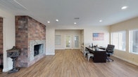 This single-floor space offers an open, versatile layout that seamlessly blends comfort with functionality. The most commanding architectural feature is the floor-to-ceiling stone and brick fireplace along one wall, providing a rustic yet refined focal point that anchors the room with warmth and character. Durable wood-look plank flooring extends throughout the space, lending a warm, cohesive aesthetic underfoot.

Recessed lighting is evenly distributed across the textured ceiling, ensuring bright, consistent illumination throughout the interior. On one side of the room, a dedicated workstation area is outfitted with a large dark desk, industrial-style metal chairs, and computer equipment, making the space well-suited for office or administrative use.

A set of double glass-panel doors at the far end of the room allows natural light to filter into the interior while providing a clear sightline to the entry area beyond. Neutral beige walls keep the atmosphere clean and inviting, complementing both the natural tones of the flooring and the earthy textures of the fireplace surround. Overall, the space strikes a balance between a welcoming, homey ambiance and a practical, well-organized work environment.