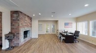 This single-floor space offers an open, versatile layout that seamlessly blends comfort with functionality. The most commanding architectural feature is the floor-to-ceiling stone and brick fireplace along one wall, providing a rustic yet refined focal point that anchors the room with warmth and character. Durable wood-look plank flooring extends throughout the space, lending a warm, cohesive aesthetic underfoot.

Recessed lighting is evenly distributed across the textured ceiling, ensuring bright, consistent illumination throughout the interior. On one side of the room, a dedicated workstation area is outfitted with a large dark desk, industrial-style metal chairs, and computer equipment, making the space well-suited for office or administrative use.

A set of double glass-panel doors at the far end of the room allows natural light to filter into the interior while providing a clear sightline to the entry area beyond. Neutral beige walls keep the atmosphere clean and inviting, complementing both the natural tones of the flooring and the earthy textures of the fireplace surround. Overall, the space strikes a balance between a welcoming, homey ambiance and a practical, well-organized work environment.