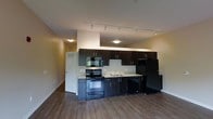 This single-floor unit showcases an open-concept design that seamlessly integrates the kitchen and living areas into one cohesive space. The kitchen is a standout feature, outfitted with sleek dark espresso cabinetry that extends to the upper and lower levels, providing generous storage. Black appliances, including a range, built-in microwave, dishwasher, and refrigerator, complement the cabinetry for a uniform, modern aesthetic. A light-toned countertop and a simple tile backsplash offer a subtle contrast to the darker elements. The kitchen sink is positioned along the counter, practical and easily accessible. Track lighting runs across the ceiling above the kitchen, ensuring the workspace is well-lit, while flush-mount fixtures illuminate the adjoining areas. The flooring throughout is a warm hardwood-style material that adds continuity across the open layout. The living area benefits from the openness of the floor plan, creating a generous and versatile space. The neutral wall tones contribute to a clean, understated atmosphere throughout the unit.