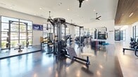 Located at 965 Duncan Perry Rd in Grand Prairie, Texas, this modern fitness center offers a generously sized, single-floor layout designed to accommodate a comprehensive range of workout activities. The space makes a strong first impression with a stylish entry foyer featuring terrazzo flooring, a slatted wood ceiling accent, globe pendant lighting, a caramel leather bench, and a bold geometric tile feature wall — setting an elevated tone before entering the main floor.

The main fitness area is defined by polished dark concrete floors, high ceilings, and expansive floor-to-ceiling steel-framed windows that flood the interior with natural light and provide open views of the surrounding outdoor environment, including a pool area. Ceiling fans are thoughtfully distributed throughout to maintain airflow across the space.

The cardio zone is well-appointed with treadmills, ellipticals, stair climbers, stationary bikes, and a rowing machine, all positioned along the window walls to maximize views. The strength training area features multi-station cable machines, free weight benches, weight racks, and a punching bag. A functional training zone includes plyo boxes and additional accessories. Wall-mounted TVs are positioned throughout the space for entertainment during workouts.