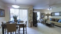 This single-floor residence offers an open-concept design that seamlessly connects the dining and living areas into one cohesive, inviting space. The dining area is anchored by a dark wood table with upholstered bar-height chairs and illuminated by a large pendant light fixture overhead. A floor-to-ceiling sliding glass door with vertical blinds floods the space with abundant natural light while providing access to an outdoor balcony, offering a bright and airy atmosphere throughout. Decorative framed artwork lines the walls, complementing the warm, neutral color palette of the interiors. A dark wood armoire serves as a striking focal point between the two spaces, flanked by sculptural accents and lush greenery that add warmth and character. The living area features plush, patterned upholstered seating arranged around a low-profile coffee table atop soft wall-to-wall carpeting. Additional framed art and a warm table lamp contribute to the relaxed yet refined ambiance. The overall aesthetic is cohesive and thoughtfully curated, blending natural textures with understated elegance to create a comfortable and functional living environment.