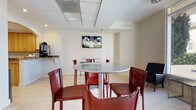 This single-floor amenity space is designed to serve as a multifunctional gathering and leisure area, finished throughout with large-format light tile flooring and neutral cream-toned walls that create a bright, clean aesthetic. The dining and social area features round tables with sleek metallic pedestals surrounded by bold red leather bar-height chairs, providing casual seating for multiple groups. Wall-mounted flat-screen televisions are positioned throughout the space for easy viewing. A console table along one wall displays reading materials beneath an additional mounted TV, offering a quieter corner for relaxation.

An arched passthrough leads to a fully equipped kitchen area outfitted with stainless steel appliances, wood cabinetry, granite countertops, and a coffee station, making the space well-suited for gatherings and events. Adjacent to the kitchen, a drop ceiling with recessed lighting adds structure to the lounge zone.

Floor-to-ceiling windows line one side of the space, flooding the interior with natural light and providing direct views of an outdoor terrace equipped with high-top patio seating and a pool beyond. A glass-enclosed fitness center is visible from the lounge area, with tall indoor plants adding a natural, welcoming element to the overall ambiance.