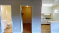 This single-floor residential space is designed with functionality and practicality in mind. The interior features light-toned walls paired with white trim throughout, creating a clean and cohesive aesthetic. Hardwood-style flooring extends across the main areas, adding warmth and continuity to the space.

A notable feature visible from the main living area is a walk-in closet, accessible through a white-trimmed doorway. Inside, the closet is equipped with a closet rod system and is warmly lit by a ceiling fixture, offering ample storage and organization potential. The walls within the closet are painted in a warm golden-yellow tone, providing a distinct contrast to the neutral palette of the main space.

Adjacent to the closet area, the kitchen is outfitted with white cabinetry along the upper and lower sections, providing generous storage. The appliances, including what appears to be a range and an under-cabinet hood, coordinate with the white cabinetry for a unified look. The kitchen space flows naturally into the surrounding areas, contributing to an open and accessible feel.

Overall, the space presents a straightforward and well-maintained interior, with practical storage solutions and a bright, neutral color scheme that offers a versatile canvas for personalization.