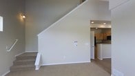 This thoughtfully designed residential space spans three floors, offering a versatile and functional layout suited for a variety of lifestyles. The main living area immediately impresses with its dramatic vaulted ceilings that rise steeply overhead, creating a strong sense of openness and volume. Soft, neutral tones on the walls and plush carpeting throughout the living space lend a warm and understated aesthetic that serves as a clean canvas for personalization.

A staircase with crisp white trim connects the floors, and its angular profile contributes to the architectural character of the main level. Recessed lighting provides even, modern illumination throughout. Just off the living area, the kitchen is visible and features light natural wood cabinetry, a dark countertop island or peninsula, and full-size appliances including a refrigerator — offering a practical and attractive space for cooking and gathering. Clean white interior doors and consistent trim work throughout reinforce a cohesive, polished design language.

The multi-floor layout provides clear separation between living, sleeping, and utility areas, making efficient use of vertical space. Overall, the space presents a clean, move-in-ready environment with well-maintained finishes, an airy atmosphere, and a layout designed to balance open communal spaces with private retreats across its three levels.