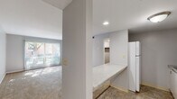 This single-floor unit is thoughtfully laid out to maximize functionality within a compact footprint. The space comprises a living room, kitchen, closet, and entrance, each serving a distinct purpose while contributing to a cohesive, efficient flow.

The living room is the heart of the unit, finished with wall-to-wall carpet that adds warmth underfoot. A large sliding glass door dressed with vertical blinds allows generous natural light to pour in, casting bright patterns across the floor and offering pleasant views of the surrounding greenery and outdoor common areas. A built-in closet with louvered bi-fold doors provides convenient storage directly within the living space.

The kitchen is efficiently designed with white upper and lower cabinetry, a speckled laminate countertop, a stainless steel sink, and a full-size white refrigerator. Tile flooring adds practicality and durability to this area. A pass-through countertop connects the kitchen to the main living area, enhancing the open feel between the two spaces.

A dedicated walk-in closet, carpeted and equipped with a full-length hanging rod and shelving, offers ample storage capacity. The entrance provides a natural transition into the unit. Throughout, the space features neutral gray walls, recessed lighting, and clean finishes that lend a simple, move-in-ready aesthetic.