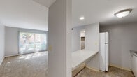 This single-floor unit is thoughtfully laid out to maximize functionality within a compact footprint. The space comprises a living room, kitchen, closet, and entrance, each serving a distinct purpose while contributing to a cohesive, efficient flow.

The living room is the heart of the unit, finished with wall-to-wall carpet that adds warmth underfoot. A large sliding glass door dressed with vertical blinds allows generous natural light to pour in, casting bright patterns across the floor and offering pleasant views of the surrounding greenery and outdoor common areas. A built-in closet with louvered bi-fold doors provides convenient storage directly within the living space.

The kitchen is efficiently designed with white upper and lower cabinetry, a speckled laminate countertop, a stainless steel sink, and a full-size white refrigerator. Tile flooring adds practicality and durability to this area. A pass-through countertop connects the kitchen to the main living area, enhancing the open feel between the two spaces.

A dedicated walk-in closet, carpeted and equipped with a full-length hanging rod and shelving, offers ample storage capacity. The entrance provides a natural transition into the unit. Throughout, the space features neutral gray walls, recessed lighting, and clean finishes that lend a simple, move-in-ready aesthetic.