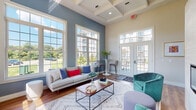 Located at 1779 Central St in Stoughton, Massachusetts, this single-floor community clubhouse offers a thoughtfully designed gathering space that balances comfort, functionality, and contemporary style. The centerpiece of the space is a spacious lounge area distinguished by soaring coffered ceilings, rich hardwood flooring, and an abundance of natural light streaming through floor-to-ceiling multi-pane windows. A sleek linear fireplace is set against a textured white shiplap accent wall, flanked by plush teal lounge chairs and a generous neutral sofa, creating an inviting and social atmosphere. French doors open to an outdoor area, further enhancing the sense of openness and connection to the surrounding landscape. The well-appointed kitchen features a large central island with a bold teal base and white quartz countertop, brown leather barstools, stainless steel appliances, white cabinetry with brass hardware, and a decorative tile backsplash — ideal for catered events or casual gatherings. Adjacent to the lounge and kitchen are two dedicated conference rooms, each bathed in natural light from oversized windows. Both rooms are furnished with long worktables and modern blue rolling chairs, providing practical settings for meetings or collaborative work. The overall design palette of soft blues, warm neutrals, and natural wood tones creates a cohesive and welcoming environment throughout the space.