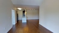 This single-floor space is defined by its open-concept layout, where the living area and kitchen share a cohesive, uninterrupted flow. Rich, dark hardwood flooring extends throughout, grounding the space with warmth and a polished aesthetic. Neutral-toned walls keep the interior bright and versatile, complementing the clean architectural lines.

The kitchen is positioned centrally, anchored by a white peninsula island with a built-in sink and stone countertop, offering both functionality and a natural division between cooking and living spaces. Dark cabinetry lines the kitchen backdrop, providing strong visual contrast against the lighter elements. A stainless steel refrigerator is visible, reinforcing the kitchen's practical design. Overhead, a pendant light fixture adds focused illumination above the island, while a track lighting system in the adjacent area ensures the living space remains well-lit.

The expansive living area provides generous room for a variety of furniture arrangements. A mirrored or glass door near the entrance adds depth and lightness to the entryway. Overall, the space presents a modern, functional, and well-appointed interior suitable for everyday living.