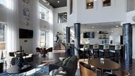 Located at 811 Spring Forest Rd in Raleigh, NC, this single-floor space makes a striking impression through its dramatic double-height ceilings and expansive windows that flood the interior with natural light, offering views of the surrounding neighborhood. The open-concept layout is anchored by distinctive mosaic-tiled columns in deep navy and black, adding a bold architectural element that contrasts beautifully with the crisp white walls and dark ceiling detail above.

A well-appointed bar area serves as a central focal point, complete with a tiled backsplash, mounted television, open shelving, and stylish bar seating upholstered in patterned green and gold fabric. Adjacent to the bar, a lounge area features contemporary dark sofas, sleek coffee tables, and a blue geometric area rug, creating a comfortable setting for relaxed conversation or casual gatherings. Smaller café-style seating clusters are positioned near the oversized windows, maximizing the abundance of natural light throughout the day.

The overall ambiance is modern and sophisticated, blending upscale design details with an inviting, social atmosphere suited for a variety of uses.