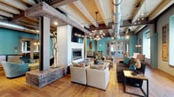The Lofts at Helmetta community space is a masterfully reimagined interior that celebrates its industrial roots while delivering a warm, stylish atmosphere. The main lounge area makes an immediate impression with its soaring exposed-beam ceilings, whitewashed wood planking overhead, and prominent silver ductwork that pays homage to the building's original character. Rich, wide-plank hardwood floors run throughout, grounding the space with warmth and texture.

At the heart of the lounge sits a striking linear fireplace set into a white surround atop an exposed brick hearth, flanked by sculptural art pieces that add a gallery-like quality. Multiple seating arrangements — including plush neutral sofas, velvet swivel chairs, and accent ottomans — are organized to encourage both intimate conversation and larger gatherings. A statement Sputnik chandelier anchors the room with dramatic effect.

Vibrant teal accent walls energize the space, providing a bold backdrop for eclectic artwork and decorative elements. Large windows along the perimeter draw in natural light, while French doors lead to private rooms ideal for meetings or smaller gatherings. A cozy dining nook features tufted leather banquette seating, industrial pendant lighting, and whitewashed brick walls, offering a distinct space for casual dining or socializing. The overall ambiance is creative, sophisticated, and unmistakably loft-inspired.
