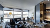 This single-floor space showcases a contemporary open-concept design that efficiently combines living, dining, and working areas into one cohesive environment. Large, floor-to-ceiling windows along the exterior wall flood the interior with abundant natural light while framing expansive urban views of the surrounding cityscape. The living area is anchored by a dark upholstered sofa paired with a sleek metal-framed coffee table, set atop a patterned area rug over warm hardwood flooring. A dedicated workspace sits adjacent to the living zone, featuring a compact desk with a wood-toned finish, a framed mirror, and a task chair — thoughtfully integrated without disrupting the flow of the space. The dining area is positioned nearby, with place settings and glassware visible, suggesting an intimate yet functional setup. The interior palette combines cool grey-blue walls with white ceilings and natural wood tones, creating a balanced, modern aesthetic. Roller shades on the windows offer light control while maintaining the clean, minimalist look throughout. The overall ambiance is stylish, urban, and efficiently designed for contemporary living.