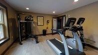 This fitness room offers a functional and organized space designed to meet a variety of exercise needs. The room is finished with light wood-style flooring that provides a clean, modern aesthetic, while the warm taupe and yellow-toned walls contribute to an inviting atmosphere. Recessed ceiling lights illuminate the space evenly, ensuring good visibility throughout.

The cardio section is anchored by a True-brand treadmill positioned prominently in the room, accompanied by an elliptical trainer nearby, both offering modern console displays for tracking workout metrics. On the opposite side, an Inspire-brand multi-function cable and weight machine with an integrated bench provides comprehensive strength training options, catering to a range of exercise routines.

A framed informational notice is mounted on the wall, likely outlining facility rules and guidelines. Safety is addressed with a mounted fire extinguisher near the exit door. A waste bin is conveniently placed nearby. A mirrored wall panel along one side enhances the sense of space. The room's single-floor layout and straightforward design make it an accessible and practical fitness facility.