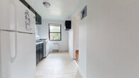 This single-floor unit offers a practical and well-organized layout, connecting several distinct living spaces through a central hallway finished with warm hardwood flooring. The living area is bright and open, featuring hardwood floors, white walls, and a large window fitted with blinds that allow ample natural light to fill the room. A radiator unit provides reliable heating throughout the space.

The kitchen is efficiently designed with dark lower and upper cabinetry that contrasts against white tile backsplash and light tile flooring. It is equipped with a gas range, a full-size refrigerator, and a stainless steel sink. A window above the counter area brings in natural light and offers views of the surrounding neighborhood.

The bedrooms continue the hardwood flooring theme, with white walls and windows that overlook greenery and neighboring brick buildings. One bedroom includes a built-in closet with a rod and shelf, offering convenient storage. Radiator heating is present in the bedrooms as well.

A hallway connects the living spaces to the bathroom, which features full wall tiling, a soaking bathtub with a shower, and a window for natural ventilation and light. The overall layout is straightforward and functional, making efficient use of the available space throughout.