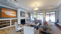 This single-floor space exudes a warm and inviting ambiance, thoughtfully designed to balance comfort with refined style. The focal point of the main lounge area is a classic white fireplace framed by built-in shelving units stocked with neatly arranged firewood, adding both a decorative and functional element to the room. Crown molding along the ceiling and crisp white architectural columns lend a sense of elegance and structure to the space.

A large pendant light fixture hangs centrally, complemented by recessed track lighting that highlights the artwork adorning the walls, including bold orange and warm-toned pieces that energize the room's palette. The neutral gray walls provide a sophisticated backdrop that ties together the furnishings seamlessly.

The seating area is generously appointed with plush sofas and accent chairs in soft gray and white geometric patterns, accented by red and orange throw pillows that echo the artwork. A rectangular coffee table anchors the arrangement atop a layered patterned rug, completing the cohesive interior design.

Large floor-to-ceiling windows and glass doors line the perimeter, inviting an abundance of natural light and offering views of an outdoor patio area with lush greenery beyond. The seamless connection between the interior lounge and the outdoor space enhances the overall sense of openness and livability.