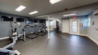 Located at 266 Providence Ave in East Providence, Rhode Island, this well-equipped fitness center is thoughtfully designed to support a full range of workout routines. The single-floor facility features durable speckled rubber flooring throughout, providing a safe and functional surface ideal for exercise. The space is divided into distinct workout zones, each serving a specific fitness purpose.

The cardio area is outfitted with multiple treadmills and an elliptical trainer, each positioned beneath wall-mounted flat-screen televisions for an engaging workout experience. Adjacent to the cardio section is a multi-functional strength training machine, offering a variety of resistance-based exercises. A dedicated free weights area includes a full rack of dumbbells and yoga/exercise mats, catering to both strength and flexibility training needs.

A standout feature of the space is the large mirrored wall accented with a bold orange stripe, which visually expands the room and supports form-checking during exercise. The space also includes a stationary spin bike positioned in front of the mirror. Bright recessed panel lighting ensures the entire area is well-illuminated, while a wall-mounted mini-split system provides climate control. Natural light filters in through windows, and the facility includes clearly marked exit access, ensuring a comfortable and safe environment for all users.