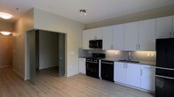 This single-floor space offers a clean, contemporary design with an open and functional layout. The kitchen is a standout feature, showcasing floor-to-ceiling white cabinetry with brushed nickel hardware, dark countertops, and a gray subway tile backsplash that adds a modern touch. A full suite of black and stainless steel appliances — including a gas range, built-in microwave, and dishwasher — provides a sleek, cohesive look. A deep sink with a modern faucet completes the kitchen workspace. Light-toned hardwood-style flooring runs throughout, lending warmth and visual continuity to the space. Neutral wall tones create a bright and versatile backdrop that complements the modern aesthetic. A sliding glass door leads to an adjacent room, adding an architectural element that enhances the open feel while maintaining the option for separation. Track lighting on the ceiling provides focused illumination in the kitchen area. The overall ambiance is modern, clean, and move-in ready, making excellent use of the available space with a well-considered design that balances style and practicality.