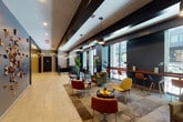 This contemporary building lobby makes a strong first impression through its thoughtful architectural design and high-end finishes. The entry foyer welcomes visitors with large-format porcelain tile flooring, bold dark ceiling beams, and dramatic sculptural pendant light fixtures that cascade from above, creating an artistic focal point. A prominent reception desk clad in richly veined marble anchors the space, positioned near a pair of stainless steel elevator doors set against a striking herringbone-patterned tile feature wall.

The lounge area flows naturally from the entry, offering a curated mix of mid-century modern seating in warm tones of mustard yellow, olive green, and rust red, arranged around low tables on a textured area rug. A long wooden counter-height bar runs along the floor-to-ceiling windows, furnished with a mix of wire and leather bar stools, providing an ideal workspace with views to the street outside. Lush indoor plants add organic warmth and texture throughout.

A dedicated office space, enclosed in glass, offers a bright and functional meeting area with its own statement chandelier and natural light. The overall ambiance balances sophistication and comfort, blending dark architectural elements with warm wood tones and vibrant furnishings to create a welcoming, design-forward environment.