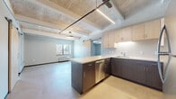 This single-floor unit showcases a thoughtfully designed interior that blends modern finishes with industrial architectural character. The open-concept kitchen is a central highlight, featuring a peninsula with a light-toned quartz countertop, dual-tone cabinetry with light maple uppers and dark wood-grain lowers, and a full suite of stainless steel appliances including a refrigerator, electric range, microwave, and dishwasher. A sliding barn door adds a contemporary touch, separating spaces with functional style.

The living area flows naturally from the kitchen, offering polished concrete flooring and an exposed concrete ceiling with structural beams that lend an industrial character. A wall-mounted HVAC unit and a window overlooking the exterior provide comfort and natural light throughout.

The carpeted bedroom areas feature the same exposed concrete ceiling aesthetic, accented by round pendant lighting and textured concrete block walls that emphasize the building's original construction. Each room is well-proportioned with dedicated windows allowing in natural light.

The bathroom is clean and functional, equipped with a vanity featuring dark blue cabinetry, a single sink, a large mirror, and a full tub-shower combination. An additional bathroom space includes a vanity with dark cabinetry and a sliding door. Polished concrete flooring runs throughout the wet areas, tying the unit's design together cohesively.
