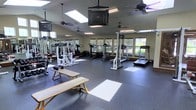 This fitness center offers a spacious, single-floor layout designed to accommodate a full range of workout activities. The room is bathed in natural light streaming through multiple skylights overhead, complemented by large windows along the walls that provide views of the surrounding greenery, creating an inviting and energizing atmosphere. Several ceiling fans are installed throughout the space, ensuring comfortable airflow during workouts. The flooring consists of durable dark rubber matting, well-suited for heavy equipment and high-impact exercise. Full-length mirrors line one wall, enhancing the sense of openness and allowing users to monitor their form. The strength training area is equipped with a multi-station cable machine, weight benches, and free weights, while the cardio section features treadmills, an elliptical trainer, and a stationary bike. Overhead, a mounted television provides entertainment during workouts. The combination of ample natural light, thoughtful ventilation, and a diverse selection of equipment makes this fitness center a functional and well-appointed space for a comprehensive exercise routine.