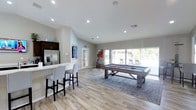 This single-floor amenity space is designed for leisure and social gathering, offering a well-appointed and inviting environment. The room is anchored by a rustic wood-framed billiards table set atop a stylish area rug, making it the centerpiece of the space. Light hardwood-style flooring flows throughout, complementing the neutral gray and white wall tones that lend the space a clean, contemporary feel.

A built-in kitchenette area lines one wall, featuring a stainless steel refrigerator with an ice and water dispenser, upper cabinetry, and a countertop bar with upholstered bar stools — ideal for entertaining. A wall-mounted flat-screen television adds to the social atmosphere.

Recessed lighting is distributed across the vaulted ceiling, ensuring the space is well-lit, while a large sliding glass door and expansive windows flood the interior with natural light and provide direct views and access to an outdoor area featuring lounge seating, suggestive of a poolside patio. The seamless indoor-outdoor connection enhances the overall ambiance, making the space feel open and airy. This recreation room is thoughtfully designed to serve as a comfortable and functional gathering hub.