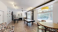 Located at 740 Smallwood Dr in Raleigh, NC, this single-floor amenity space at Wedgwood offers a well-appointed layout designed for both leisure and community gathering. The entry foyer greets visitors with large glass doors framed by views of mature surrounding trees, complemented by warm wood-toned flooring and classic décor including a decorative mirror and credenza.

The expansive lobby area flows openly from the entrance, featuring wide-plank wood-look tile flooring arranged in a parquet pattern, white wainscoting with a dark accent rail, and structural columns that define the space without interrupting its openness. Comfortable seating — including a leather sofa and vibrant mustard-yellow accent chairs paired with a glass coffee table — creates an inviting lounge atmosphere. A handsome wooden display cabinet adds a classic touch.

The game room is a standout highlight, filled with natural light from large windows overlooking lush greenery. It features an air hockey table, foosball table, a ceiling fan with track lighting, and a casual dining nook with a round wood table and pendant light. Retro wall signage adds a playful, nostalgic character.

Additional rooms, finished with neutral carpet tile flooring and clean white walls, offer flexible utility space accessible via paneled white doors, rounding out this thoughtfully designed community amenity.