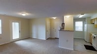 This single-floor space features an open-concept layout that connects the living area to the kitchen via a raised pass-through counter, creating a functional and social flow between spaces. The living area is finished with wall-to-wall neutral carpeting, contributing to a warm and comfortable atmosphere throughout.

The entryway includes two white paneled doors — likely a front entry and a closet — providing convenient storage and a defined point of arrival. The pass-through counter offers a casual serving or workspace that bridges the kitchen and living area, with a small decorative ledge above adding subtle architectural interest.

Walls throughout are painted in a soft, neutral tone, complementing the light carpeting and white trim work. Flush-mount ceiling light fixtures provide consistent overhead lighting across the space. The textured ceiling adds a modest visual layer to the otherwise clean and straightforward interior design.

Overall, the space presents a clean, functional, and low-maintenance interior well-suited for everyday living, with a cohesive neutral palette and practical layout that maximizes usability across the open floor plan.