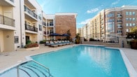 This multi-story residential community is built around a striking courtyard-style layout, with classic brick and neutral-toned exteriors complemented by black wrought-iron railings and private balconies. The centerpiece of the outdoor area is a generously sized swimming pool with crystal-clear blue water, surrounded by a spacious tiled deck. Cushioned lounge chairs and navy blue market umbrellas are arranged poolside, creating a relaxed, resort-like atmosphere. Lush landscaping and colorful plantings line the perimeter, while decorative iron fencing defines the space and adds architectural interest.

The interior common areas are finished to a high standard. A welcoming lobby-style corridor features dark-framed glass doors, wood-look flooring, and clean white walls that create a modern, transitional aesthetic. The clubhouse lounge is a well-appointed gathering space with warm leather seating, a marble-top coffee table, bold artwork, and a large wall-mounted television framed by dark accent beams and a contemporary chandelier overhead. An adjoining bar area showcases a rich dark granite countertop, upholstered bar stools with nailhead trim, decorative vessels, and tiled wall accents — all contributing to a sophisticated, hotel-inspired ambiance throughout the shared spaces.