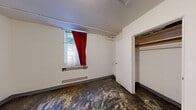 This single-floor space consists of one room with a straightforward, utilitarian layout. The walls are painted white throughout, providing a clean, neutral backdrop, while the textured ceiling adds a subtle architectural detail. An exposed pipe and track system runs along the upper portion of the walls and ceiling, offering both a functional and industrial aesthetic element.

The room features a built-in closet with open shelving, providing storage potential. A baseboard heater unit is positioned beneath the primary window, which is dressed with a sheer white curtain layered with a bold red drape, allowing for both natural light and privacy. The window frames a view of outdoor greenery, bringing a touch of the natural environment indoors.

The flooring shows significant wear and distressing across its surface, indicating the space is in need of renovation or restoration. Standard electrical outlets are placed at intervals along the walls, and a door with a lever-style handle provides access to the room. The overall condition of the space suggests it is currently vacant and presents an opportunity for renovation, making it well-suited for those looking to customize or restore the interior to a desired finish.