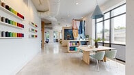 Located at 1647 E Colonial Drive in Orlando, Florida, this thoughtfully designed single-story commercial space offers a generous, open-plan layout with a bright and contemporary aesthetic. Polished concrete floors extend throughout, reflecting natural light that pours in through expansive floor-to-ceiling steel-framed windows, providing unobstructed views of the surrounding streetscape.

The interior is divided into distinct functional zones that flow seamlessly into one another. A wide, welcoming corridor greets visitors at the entrance, flanked by light wood cabinetry and textured stone columns that lend warmth and structure to the space. Further in, a collaborative meeting area features a solid wood table paired with soft sage-green chairs beneath a statement blue cone pendant light, with open shelving displaying colorful books and creative materials nearby.

A lounge area anchors another portion of the space, furnished with modular gray seating arranged on a patterned area rug, accented by bold fuchsia pendant lights that add a playful, energetic touch. Exposed HVAC ductwork runs along the white ceilings, contributing to the space's modern industrial character. Artwork and textile displays are integrated throughout, enhancing the creative atmosphere. Private rooms with dark-framed glass partitions and vibrant accent lighting offer additional versatility within this dynamic, well-appointed commercial environment.