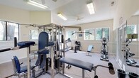 This fitness center offers a bright and welcoming environment designed to support a well-rounded workout routine. The space features vaulted ceilings that create an open, airy atmosphere, complemented by ceiling fans that promote comfortable airflow during exercise. Large windows line the walls, flooding the room with natural light and providing pleasant views of the surrounding greenery outdoors.

The facility is comprehensively equipped for both cardiovascular and strength training. Cardio equipment includes treadmills, a recumbent stationary bike, and an elliptical machine, providing ample options for aerobic exercise. For strength training, a multi-station cable machine and weight system serve as the centerpiece, accompanied by a weight bench and a selection of free weights, including dumbbells.

Full-length wall mirrors enhance the sense of space while allowing users to monitor their form during workouts. The neutral, warm-toned walls and light carpeted flooring contribute to a clean and comfortable aesthetic throughout. French doors provide access to the exterior, adding convenience and further connecting the interior to the outdoors. The layout is thoughtfully organized to maximize usable floor space while keeping equipment easily accessible.