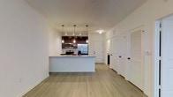 This single-floor residence showcases a thoughtfully designed open-concept layout that seamlessly connects the kitchen and living areas. The kitchen serves as the centerpiece of the space, featuring dark espresso cabinetry, a granite-topped peninsula island with an undermount sink, and a full suite of stainless steel appliances including a French door refrigerator, stainless range, and over-the-range microwave. Three elegant pendant lights hang above the island, providing focused task lighting and adding a contemporary decorative touch. The crisp white walls and smooth ceilings create a bright, airy atmosphere throughout, while light wood-style luxury vinyl flooring runs continuously across the space, lending warmth and cohesion to the overall design. A hallway visible from the main living area leads to additional rooms, flanked by white panel doors — including a louvered closet door — that contribute to the clean, modern aesthetic. The open sightlines between the kitchen and living areas make the space feel expansive and welcoming, ideal for both everyday living and entertaining. The combination of modern finishes, neutral tones, and functional design elements results in a polished and inviting interior.