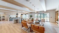 Located at 2009 Main St in Stratford, Connecticut, this generously sized single-floor community space combines contemporary design with warm, welcoming details throughout. The open-plan kitchen is a standout feature, showcasing light wood cabinetry, a white tile backsplash, a large island with seating for multiple bar stools, and a series of sleek pendant lights overhead. Built-in open shelving adds both functionality and visual interest to the space.

The adjacent living room exudes comfort and sophistication, highlighted by exposed wood ceiling beams, a stacked stone fireplace with a mounted television, and a built-in reading nook with shelving. Rich hardwood-style flooring flows seamlessly throughout, tying the interior together with a cohesive, natural aesthetic.

Large floor-to-ceiling windows and glass doors flood the interior with natural light and offer direct views of the landscaped outdoor courtyard. Outside, the expansive grounds feature lush green lawn, mature trees, curated garden beds, and multiple seating areas including Adirondack chairs and lounge furniture. A white pergola anchors an outdoor dining area, complementing the crisp white exterior of the multi-story building with its private balconies and bold navy-framed windows.