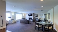 This single-floor space offers a thoughtfully designed open-plan layout that seamlessly integrates a lounge and dining area within one expansive room. The living area is anchored by a classic fireplace with a wall-mounted flat-screen television above it, creating a natural focal point for the space. Comfortable dark upholstered sofas and armchairs are arranged to encourage conversation and relaxation, complemented by a light-toned accent chair that adds visual contrast. Recessed lighting throughout the ceiling provides consistent, even illumination, while large windows along the perimeter flood the interior with natural light and lend an open, airy feel. The carpeted flooring in a neutral tone ties the room together and adds warmth underfoot. The dining area features a sleek glass-top table surrounded by dark chairs, offering a functional and stylish setting for meals or gatherings. A decorative mirror and accent pieces adorn the walls near the fireplace, adding a refined touch to the overall aesthetic. The neutral color palette of warm beige walls, grey tones, and dark furnishings creates a cohesive, modern ambiance throughout the space.