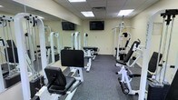 The Copper Creek fitness center is a dedicated workout facility designed to support a well-rounded exercise routine. The room is outfitted with multiple strength training machines, including cable systems, a leg press, and a leg extension/curl machine, providing residents with a comprehensive range of resistance training options. Large wall-to-wall mirrors line one side of the room, enhancing the sense of space while allowing users to monitor their form during workouts. The flooring features durable, dark carpet suited for a fitness environment. Overhead, a drop ceiling with flush-mounted lighting panels keeps the space bright and well-lit. Two flat-screen televisions are mounted on the walls, offering entertainment while exercising. The neutral, light-colored walls contribute to a clean and focused atmosphere. The layout is thoughtfully arranged to allow comfortable movement between equipment, making the fitness center a practical and functional amenity for residents of Copper Creek.