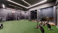 This single-floor fitness facility is designed with a contemporary aesthetic, characterized by dark charcoal walls, artificial turf flooring, and bold linear LED lighting embedded in the ceilings. The lighting system creates a dynamic, high-energy atmosphere across the space while providing even illumination throughout each zone.

The main workout area is generously proportioned, offering ample room for both strength training and functional fitness. Equipment includes a power rack, adjustable benches, a dumbbell rack, and a rowing machine, all arranged to maximize usable floor space. Motivational signage, including recognizable athletic branding, accents the walls and contributes to an energizing environment.

A dedicated cardio section houses multiple treadmills equipped with individual digital screens and stair climbers, set against mirrored walls that visually expand the room. The artificial turf flooring extends throughout, maintaining a cohesive design language.

Two enclosed glass-walled private studios provide separate spaces for cycling or focused training sessions. These rooms feature hard flooring, independent lighting, and full glass panel walls that maintain visual connectivity to the main floor.

A light-toned corridor with large glass entry doors connects the facility to the broader building, offering a clean and welcoming transition into the space.