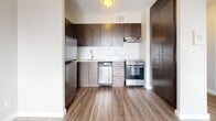 This thoughtfully designed one-bedroom, one-bathroom unit offers an efficient and comfortable single-floor layout. Throughout the space, warm wood-look laminate flooring and neutral-toned walls create a clean, contemporary aesthetic.

The kitchen is a functional standout, featuring rich dark wood cabinetry that extends to the ceiling for ample storage, a white tile backsplash, light stone countertops, and a full suite of stainless steel appliances including a refrigerator, dishwasher, and range with oven. An overhead exhaust hood and ceiling-mounted light fixture complete the practical setup.

The living room is an open, airy space with smooth white walls and generous floor area, offering a versatile blank canvas for a variety of furniture arrangements. It flows seamlessly into the rest of the unit through a connecting hallway.

The bedroom is a well-proportioned retreat, benefiting from a large window that brings in natural light and frames outdoor views. A baseboard heating unit runs along the exterior wall, and a flush ceiling fixture provides overhead lighting.

The bathroom is finished in a modern style with large-format white floor tiles, a dark wood vanity with a white countertop, an undermount sink, sleek chrome fixtures, and a full-width mirror. A dedicated closet offers additional storage within the unit.