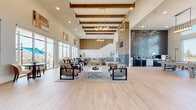 This single-story clubhouse is a masterfully designed amenity space that blends contemporary style with warm, inviting materials. The expansive main lounge commands attention with soaring ceilings adorned with exposed wood beams and statement gold-toned chandeliers, while light hardwood-style flooring grounds the open layout. Multiple seating arrangements — featuring leather and upholstered chairs, sectional sofas, and accent ottomans — are thoughtfully positioned throughout, creating distinct social zones within the generous open floor plan. A dramatic coffee bar runs along one wall, finished with a textured, three-dimensional tile backsplash and equipped with draft taps and professional-grade brewing equipment. Floor-to-ceiling windows and sliding glass doors flood the interior with natural light and provide seamless views of the outdoor pool deck, complete with lounge seating and vibrant umbrella shading. A dedicated co-working area offers workstations with computer access, surrounded by large windows overlooking the exterior. Intimate booth-style seating nooks lined with wood-textured wall panels provide quieter spots for focused work or conversation. The entry area features large-format tile flooring, curated wall art, a built-in upholstered bench, and a fully equipped mail and package locker system, completing this thoughtfully appointed community hub.