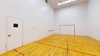 This single-room racquetball court offers a well-appointed, regulation-style playing environment designed for serious recreational use. The court is defined by its polished hardwood floor, finished in a warm natural tone and marked with multiple sets of court lines in red, green, and blue — accommodating a variety of game variations and skill levels.

The surrounding walls are smooth and painted white, providing the consistent, high-contrast surface necessary for gameplay. A clearly marked front wall features a large service box outline, while a horizontal "short line" is visible on the side wall, delineating the key zones of play. The rear wall includes a standard entry door with a small viewing window, allowing observation from outside the court.

The ceiling soars well above the floor, contributing to the open, expansive feel of the space and ensuring unrestricted movement during play. Overhead, skylight panels and recessed ventilation provide natural light supplemented by interior lighting, keeping the court bright and well-ventilated. The overall layout is clean, functional, and purpose-built, making this an excellent facility for both casual players and competitive athletes looking for a dedicated racquetball space.